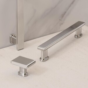 May include: A set of three brushed nickel cabinet hardware pieces. The set includes a rectangular knob, a long bar pull, and a vertical handle. The hardware has a modern, minimalist design and a matte finish.