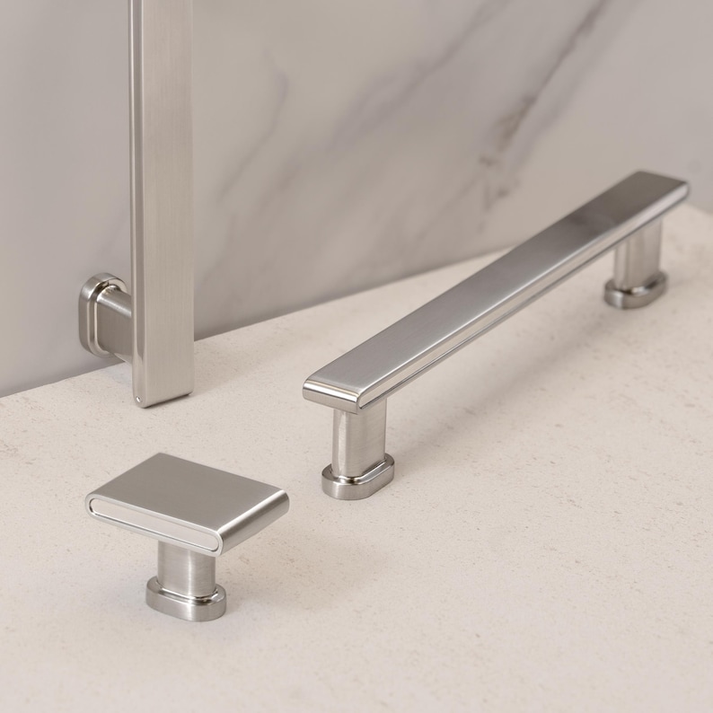 May include: A set of three brushed nickel cabinet hardware pieces. The set includes a rectangular knob, a long bar pull, and a vertical handle. The hardware has a modern, minimalist design and a matte finish.