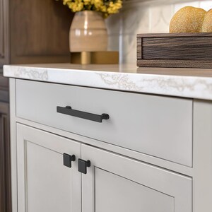 May include: A close-up of a light gray cabinet with black hardware. The cabinet has a long, rectangular handle on the drawer and square knobs on the doors. The countertop is white with gray veining.