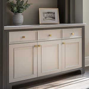 May include: A built-in cabinet with a light gray finish and gold hardware. The cabinet has three doors and three drawers. A framed black and white photo and a decorative vase with greenery sit on top.