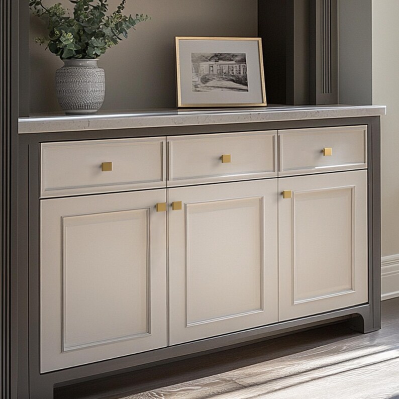 May include: A built-in cabinet with a light gray finish and gold hardware. The cabinet has three doors and three drawers. A framed black and white photo and a decorative vase with greenery sit on top.