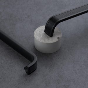 May include: Two matte black metal cabinet pulls with a ribbed, linear texture on the curved ends. The handles feature a long, smooth central bar and are positioned near a small, cylindrical gray concrete block on a dark gray textured surface.