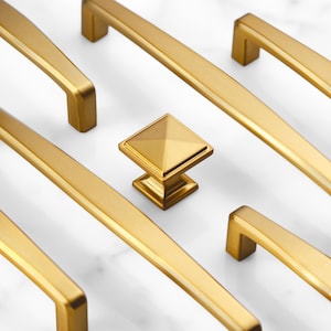 May include: A collection of brushed gold cabinet hardware including several long, rectangular bar pulls with squared ends and a single square-shaped cabinet knob with a pyramid-style top, all featuring a smooth, metallic finish.