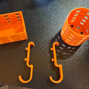 May include: Four orange 3D printed items: a small box with a grid pattern, a cylindrical container with a grid pattern, and two hooks with a J-shape.