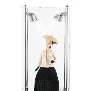 May include: A clear acrylic display case with a black base and two spotlights. A doll wearing a white blouse, black skirt, and a straw hat is displayed inside the case.