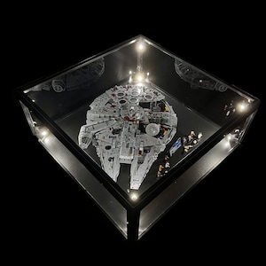 May include: A large, detailed LEGO model of the Millennium Falcon spaceship, displayed in a clear glass case with black trim and LED lights. The model is lit from above, highlighting its intricate details.