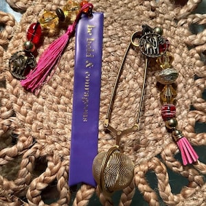 Può includere: Un infusore da tè color oro con una sfera a rete e una spilla decorativa con un ciondolo a forma di elefante. Un'etichetta viola con il testo "Be bold & courageous" è attaccata. L'infusore è adornato con perline e un nappa rosa.