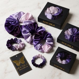 May include: A collection of purple and lavender ombre hair scrunchies in various sizes, three black square gift boxes with circular cutouts, and a rectangular black tag featuring a gold butterfly graphic and the text NOCHKA 100% Pure Mulberry Silk.