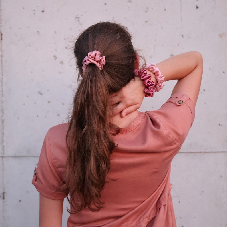May include: A woman with long brown hair wearing a pink shirt and pink scrunchies in her hair.