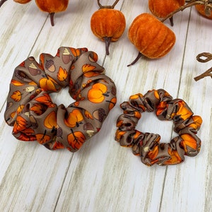 May include: Two brown and orange fabric scrunchies with a fall-themed print of pumpkins, acorns, and leaves.