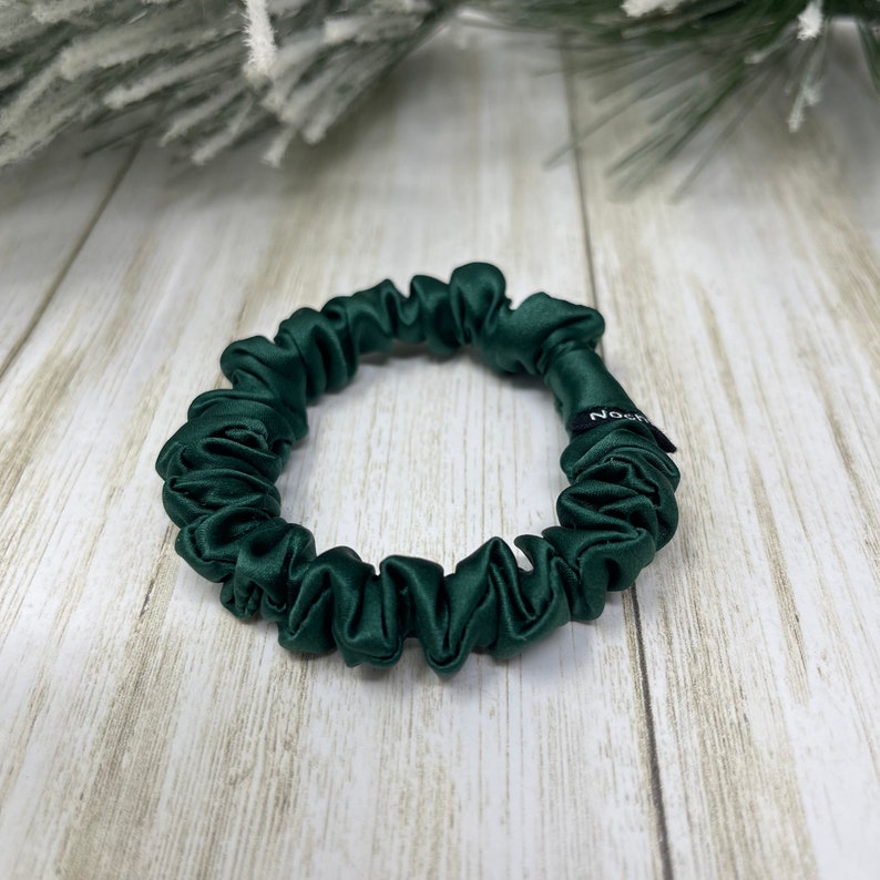 May include: A dark green satin scrunchie with a black label that says "NOSH" on a white wooden surface.