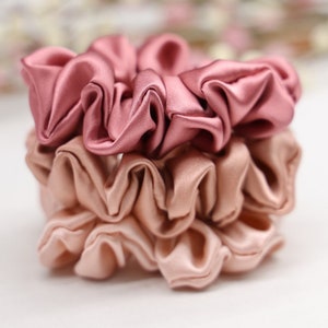 May include: Two stacked scrunchies made of soft, silky fabric. The top scrunchie is a deep rose color, and the bottom scrunchie is a light blush color.