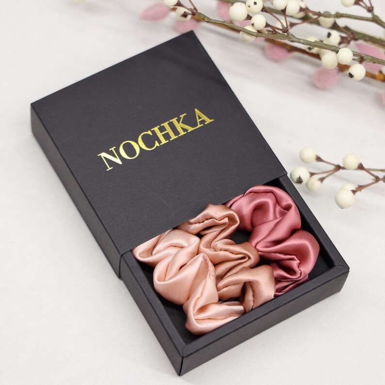 May include: Two satin scrunchies in a black box with the word "NOCHKA" printed in gold. The scrunchies are in shades of pink.