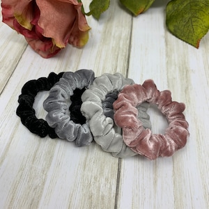 May include: Five scrunchies in different colors: black, gray, light gray, and pink. The scrunchies are made of soft, velvety fabric.