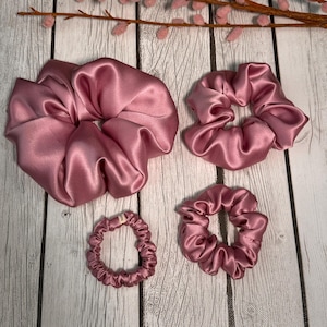 May include: Four pink satin scrunchies of different sizes. The largest scrunchie is in the top left corner, the second largest is in the top right corner, the smallest is in the bottom left corner, and the medium-sized scrunchie is in the bottom right corner.