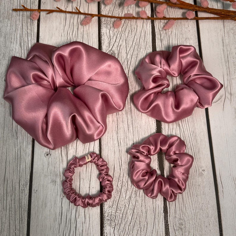 May include: Four pink satin scrunchies of different sizes. The largest scrunchie is in the top left corner, the second largest is in the top right corner, the smallest is in the bottom left corner, and the medium-sized scrunchie is in the bottom right corner.
