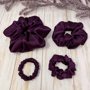 May include: Four purple satin scrunchies in different sizes. The scrunchies are on a white wooden surface.