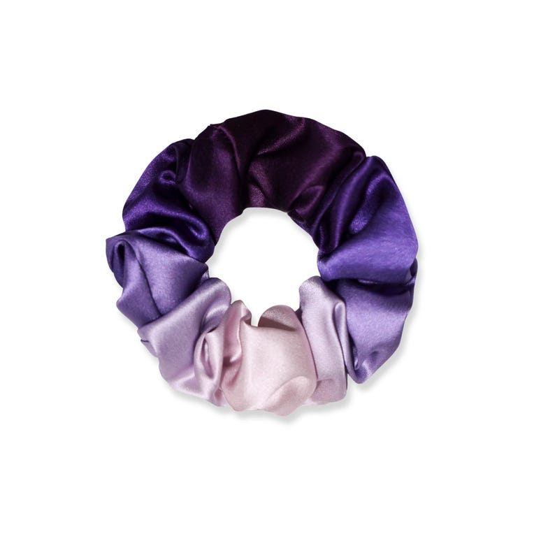 May include: A circular hair scrunchie made of smooth, shiny fabric featuring a gradient color transition. The color shifts from a deep, dark purple at the top to a lighter, soft lavender and pale pink shade at the bottom, creating a seamless ombre effect.