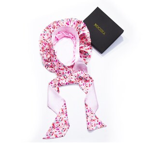 Op de afbeelding: Een roze satijnen slaapmuts met een kleurrijk splatterprint en een bijpassende stropdas. De muts is ontworpen om het haar te beschermen tijdens het slapen.