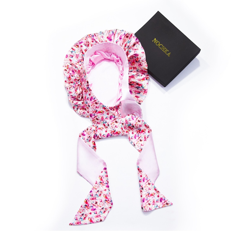 Op de afbeelding: Een roze satijnen slaapmuts met een kleurrijk splatterprint en een bijpassende stropdas. De muts is ontworpen om het haar te beschermen tijdens het slapen.