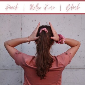 May include: A person with long brown hair wearing a pink shirt and two pink scrunchies in their hair. The scrunchies are in the colors "Punch", "Mellow Rose", and "Blush".