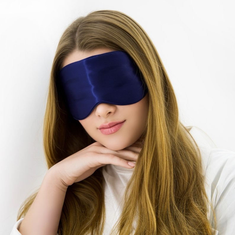 May include: A woman with long blonde hair is wearing a blue silk sleep mask. She is looking down and has her hands resting on her chin.