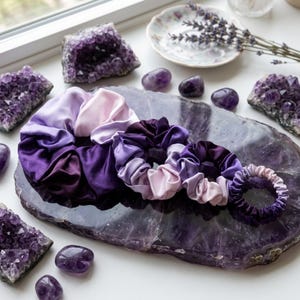 May include: A set of four hair scrunchies in varying sizes, featuring a smooth, satin-like fabric with an ombre gradient transitioning from deep purple to light lavender. The scrunchies are arranged in a row, showcasing their gathered, pleated texture.