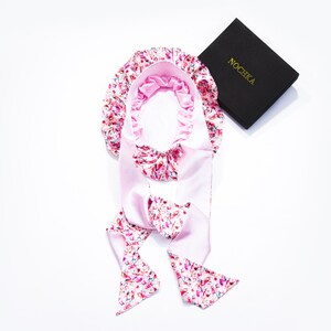 Op de afbeelding: Een roze satijnen slaapmuts met een kleurrijk bloemenprint en bijpassende banden. De muts is ontworpen om het haar te beschermen tijdens het slapen. De merknaam "NOCHKA" staat gedrukt op een zwarte doos.