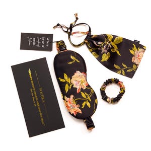 Op de afbeelding: Een zwart zijden slaapmasker met een bloemenpatroon, een bijpassende zwarte zijden tas en een zwart zijden scrunchie. Het slaapmasker heeft een gouden elastiek en de tas heeft een trekkoord. De tekst op de kaart luidt "NOCHKA "Waar luxe zijden perfectie ontmoet" 100% Pure Mulberry Silk."