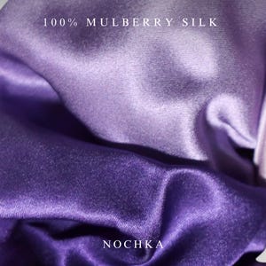 May include: A piece of fabric with a smooth, lustrous finish in shades of purple and lavender. The text 100% MULBERRY SILK is printed at the top, and the word NOCHKA is printed at the bottom. The material is draped to show its reflective surface.