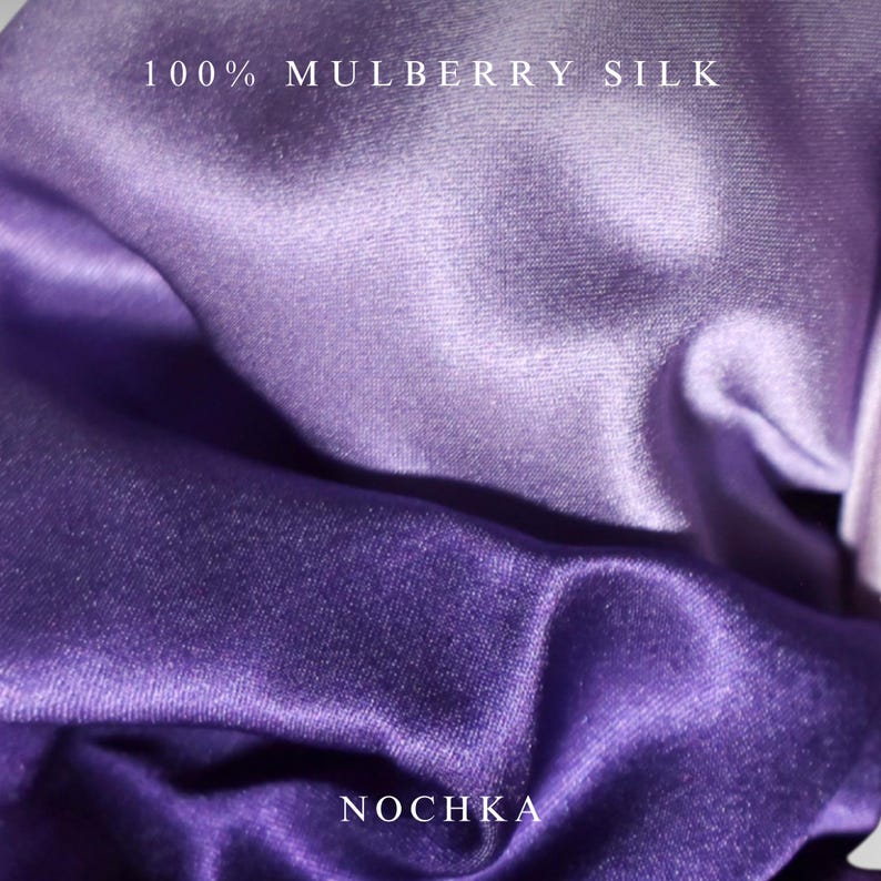 May include: A piece of fabric with a smooth, lustrous finish in shades of purple and lavender. The text 100% MULBERRY SILK is printed at the top, and the word NOCHKA is printed at the bottom. The material is draped to show its reflective surface.