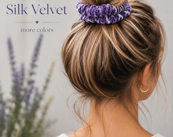 May include: A purple velvet hair scrunchie with a textured, gathered fabric design. The text 100% Silk Velvet and more colors is visible in the upper left corner. The scrunchie is positioned around a bun hairstyle with blonde highlights.