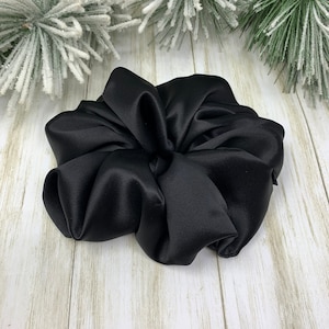 K&ouml;nnte beinhalten: Ein schwarzes Satin-Haargummi, ein Haargummi aus weichem, glattem Stoff. Das Haargummi ist in der Mitte zusammengezogen und gekr&auml;uselt, wodurch eine gro&szlig;e, fluffige Form entsteht.