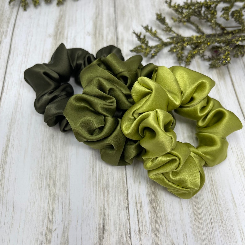 May include: Three green satin scrunchies in different shades of green. The scrunchies are arranged in a pile on a light wood surface.