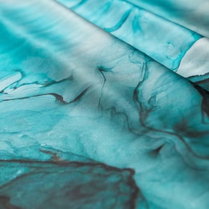 May include: Close-up of a teal blue fabric with a marbled pattern. The fabric has a soft, silky texture and is perfect for creating unique and stylish projects.
