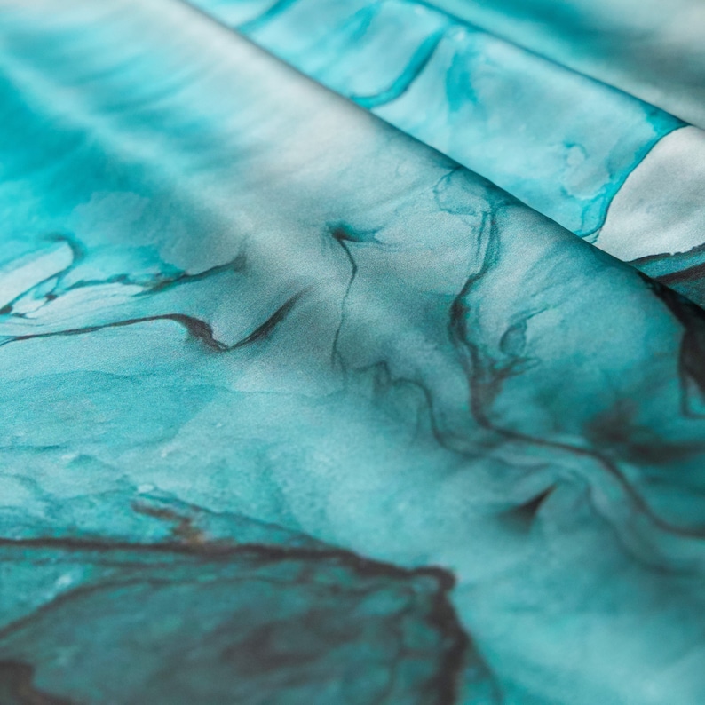 May include: Close-up of a teal blue fabric with a marbled pattern. The fabric has a soft, silky texture and is perfect for creating unique and stylish projects.
