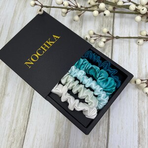 K&ouml;nnte beinhalten: Ein Set aus f&uuml;nf Satin-Haargummis in Blaut&ouml;nen und Wei&szlig;, angeordnet in einer schwarzen Box mit dem Wort "NOCHIKA" auf dem Deckel.