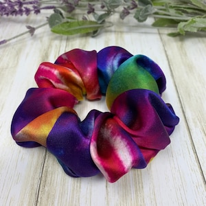 Puede incluir: Un scrunchie de color tie-dye hecho de tela de satén. El scrunchie presenta tonos de morado, rosa, naranja, amarillo, verde y azul.