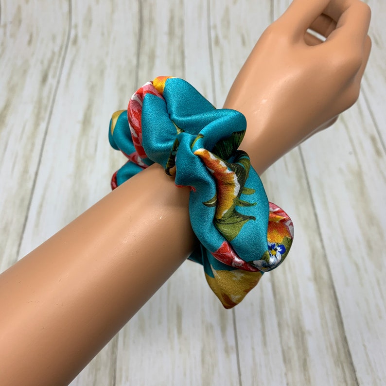 May include: A teal blue and floral patterned scrunchie. The scrunchie is made of satin fabric and has a gathered design.