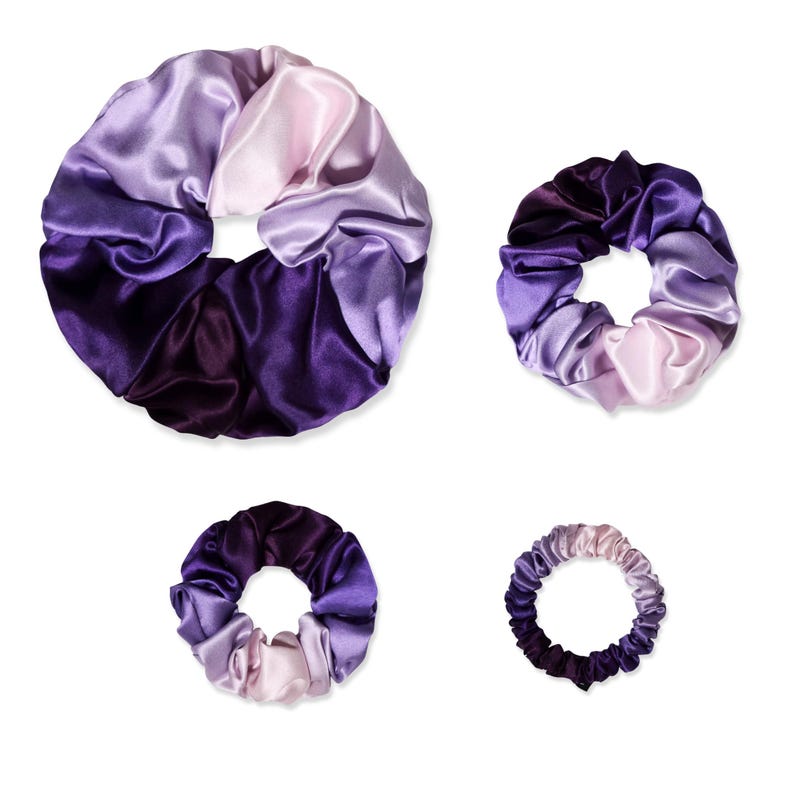 May include: Four circular hair scrunchies of varying sizes, each featuring a smooth, shiny fabric with a gradient color pattern transitioning from deep dark purple to light lavender and pale pink, arranged against a plain white background.