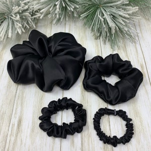K&ouml;nnte beinhalten: Vier schwarze Satin-Haargummis in verschiedenen Gr&ouml;&szlig;en. Das gr&ouml;&szlig;te Haargummi ist ein gro&szlig;es, lockeres Dutt-Haargummi. Die anderen drei sind kleiner, traditionellere Haargummis.