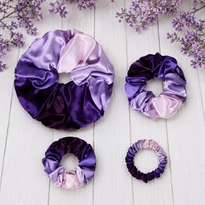 May include: Four circular hair scrunchies of varying sizes, each featuring a smooth, shiny fabric with a gradient color pattern transitioning from deep dark purple to light lavender. The scrunchies are arranged in a cluster on a white surface.