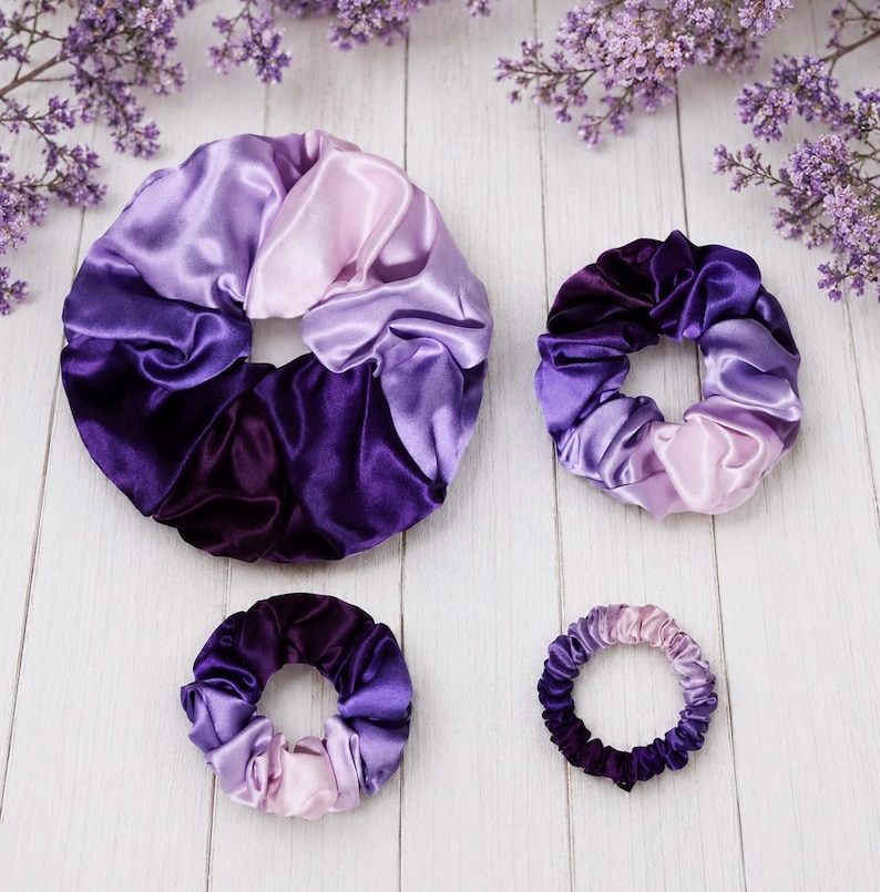May include: Four circular hair scrunchies of varying sizes, each featuring a smooth, shiny fabric with a gradient color pattern transitioning from deep dark purple to light lavender. The scrunchies are arranged in a cluster on a white surface.