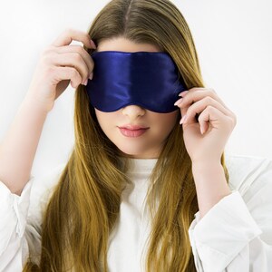 May include: A woman wearing a blue satin sleep mask over her eyes. She is wearing a white shirt and has long, brown hair.