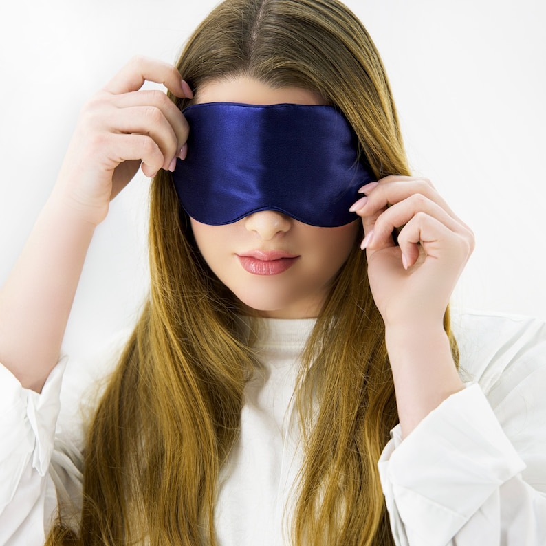 May include: A woman wearing a blue satin sleep mask over her eyes. She is wearing a white shirt and has long, brown hair.