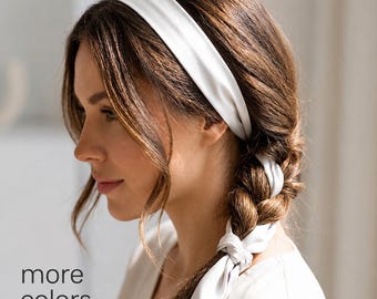 Puede incluir: Una diadema de tela blanca lisa que se lleva sobre la frente, con una cola larga que se integra en una trenza. El material liso y mate envuelve la cabeza, sujetando el cabello en un estilo sencillo y funcional con un nudo en el extremo.