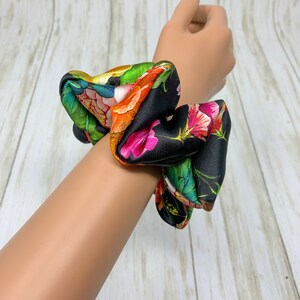 May include: A black scrunchie with a floral print featuring pink, orange, green, and white flowers. The scrunchie is made of satin fabric and is tied in a knot.