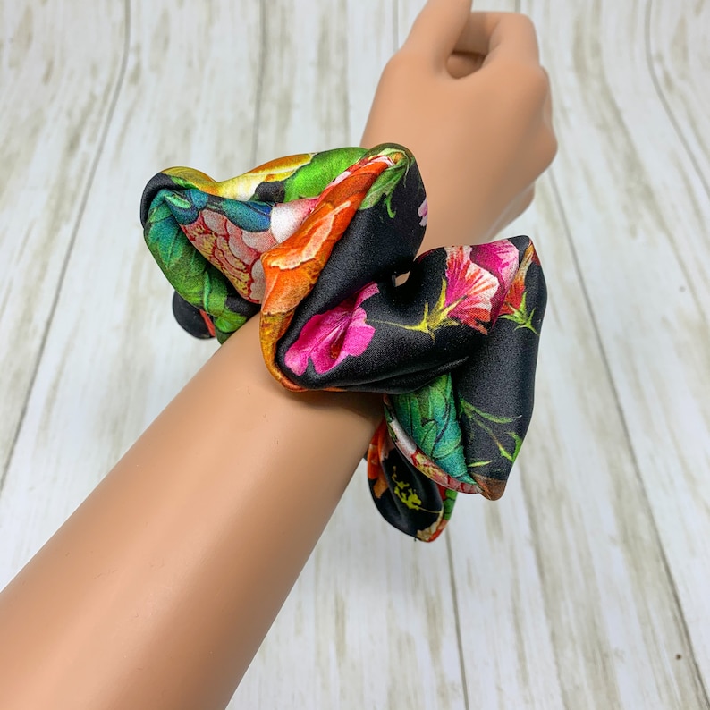 May include: A black scrunchie with a floral print featuring pink, orange, green, and white flowers. The scrunchie is made of satin fabric and is tied in a knot.