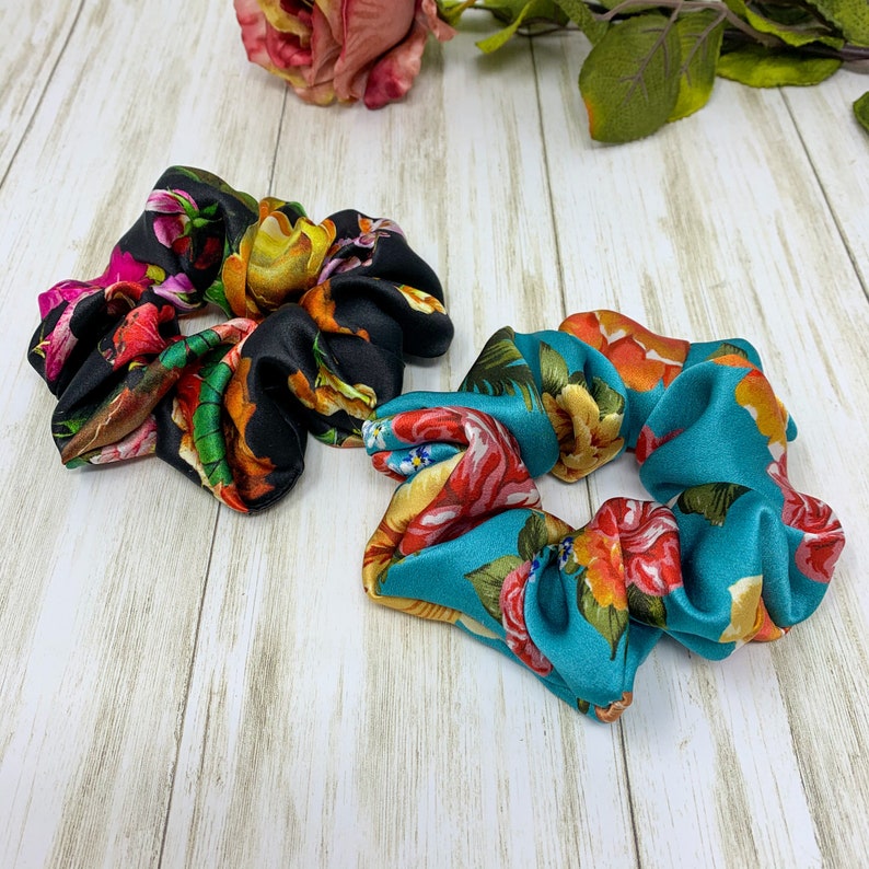 May include: Two floral print hair scrunchies. One is black with orange, yellow, and pink flowers. The other is teal with orange and yellow flowers.