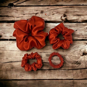 Pode incluir: Quatro scrunchies de cetim vermelho de diferentes tamanhos. O scrunchie maior está no centro da imagem, com dois scrunchies menores à direita e um scrunchie menor à esquerda.